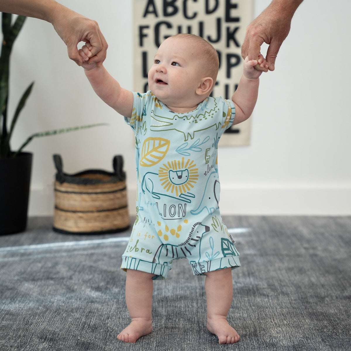 A baby wearing a light blue short-sleeve romper with a playful animal and nature print stands barefoot on a gray rug, holding hands with adult hands reaching up to support them. The romper features a lion, zebra, leaf, and the word 'LION' in yellow and green.