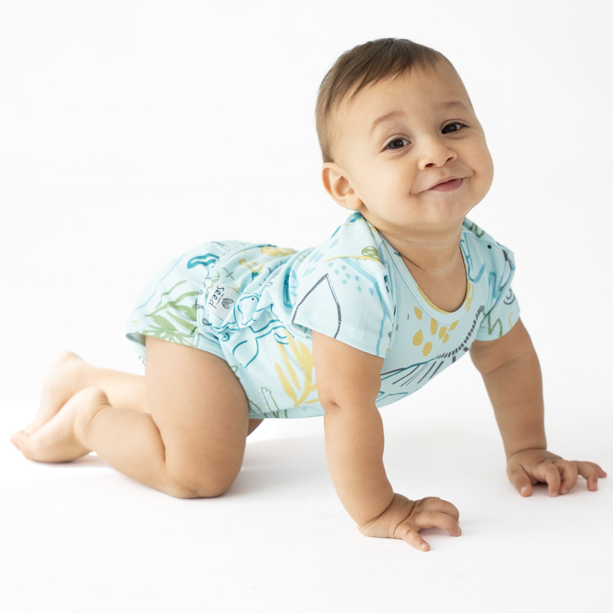 A baby crawling on a white studio floor wearing a light blue short-sleeve romper with a tropical leaf pattern in green and yellow.
