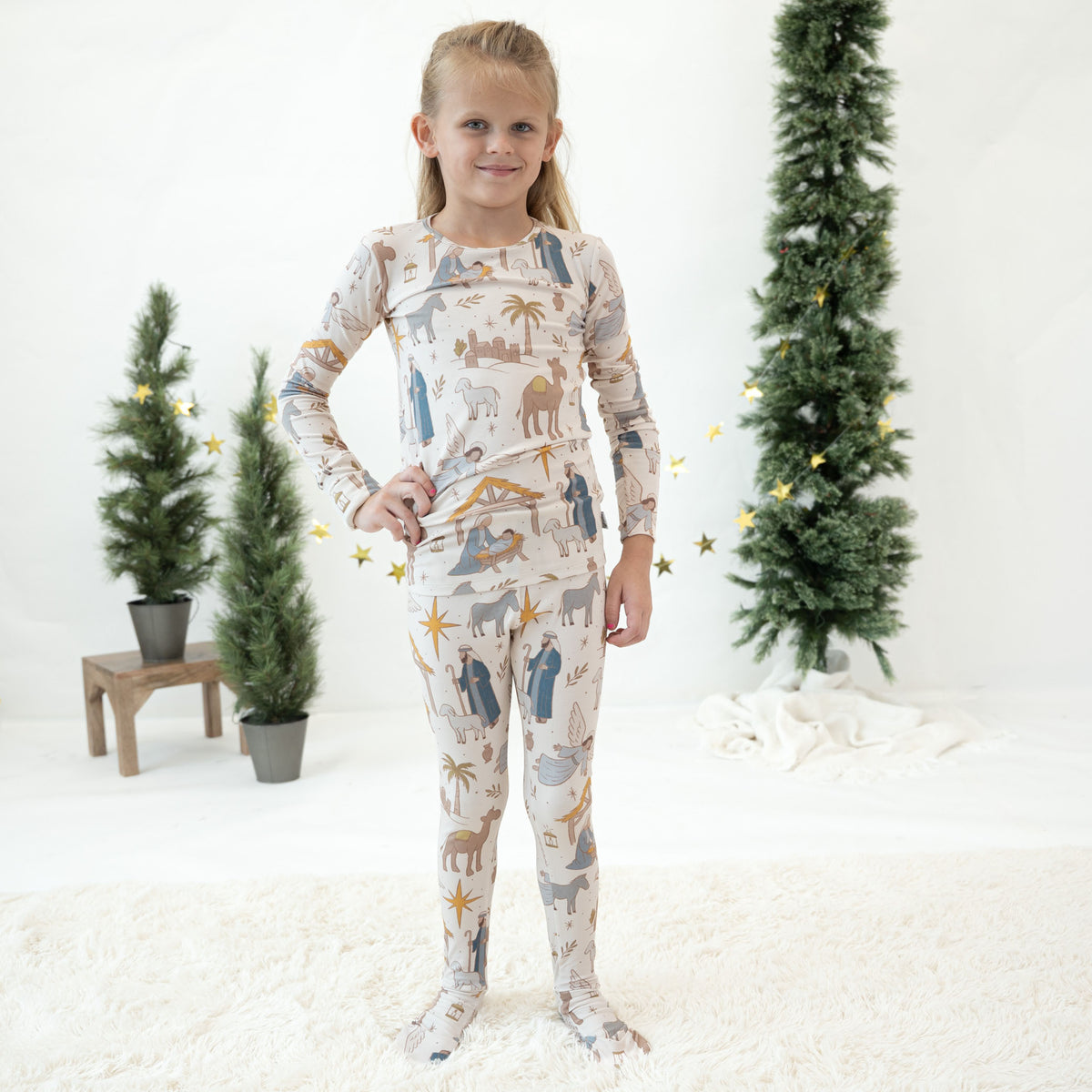 A young girl stands on a white fluffy rug wearing a long-sleeve and matching leggings pajama set with a Christmas nativity print featuring animals, stars, and palm trees. The background is a bright white studio setting decorated with potted Christmas trees and gold star accents.