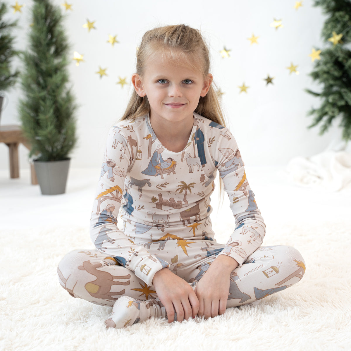 A young girl with blonde hair sitting cross-legged on a white fluffy rug, wearing a long-sleeve pajama top and matching pants with a safari-themed print of animals and palm trees. The background is a bright white room decorated with gold star lights and potted trees.