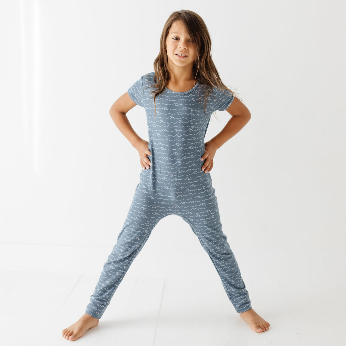 Kid stand on white floor wearing blue romper with waves. 