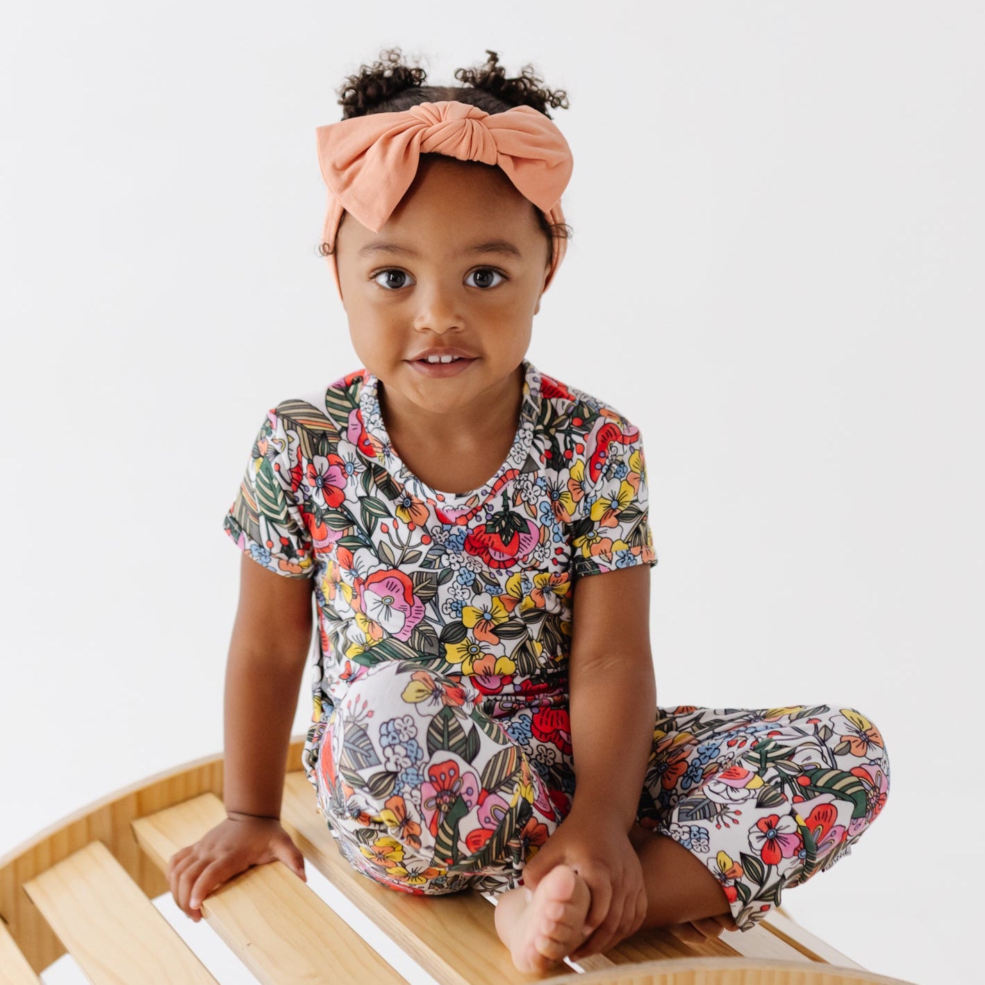 A toddler girl wearing a colorful floral print short-sleeve top and matching pants, along with a peach headband, sitting on a curved wooden bench against a plain white background.