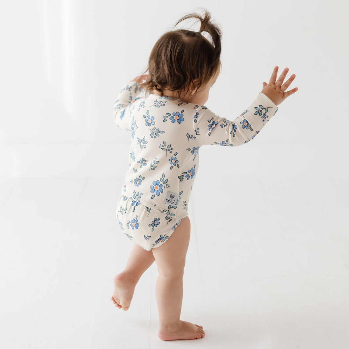 A baby stands on a white floor wearing a cream onesie with blue flowers on it. 