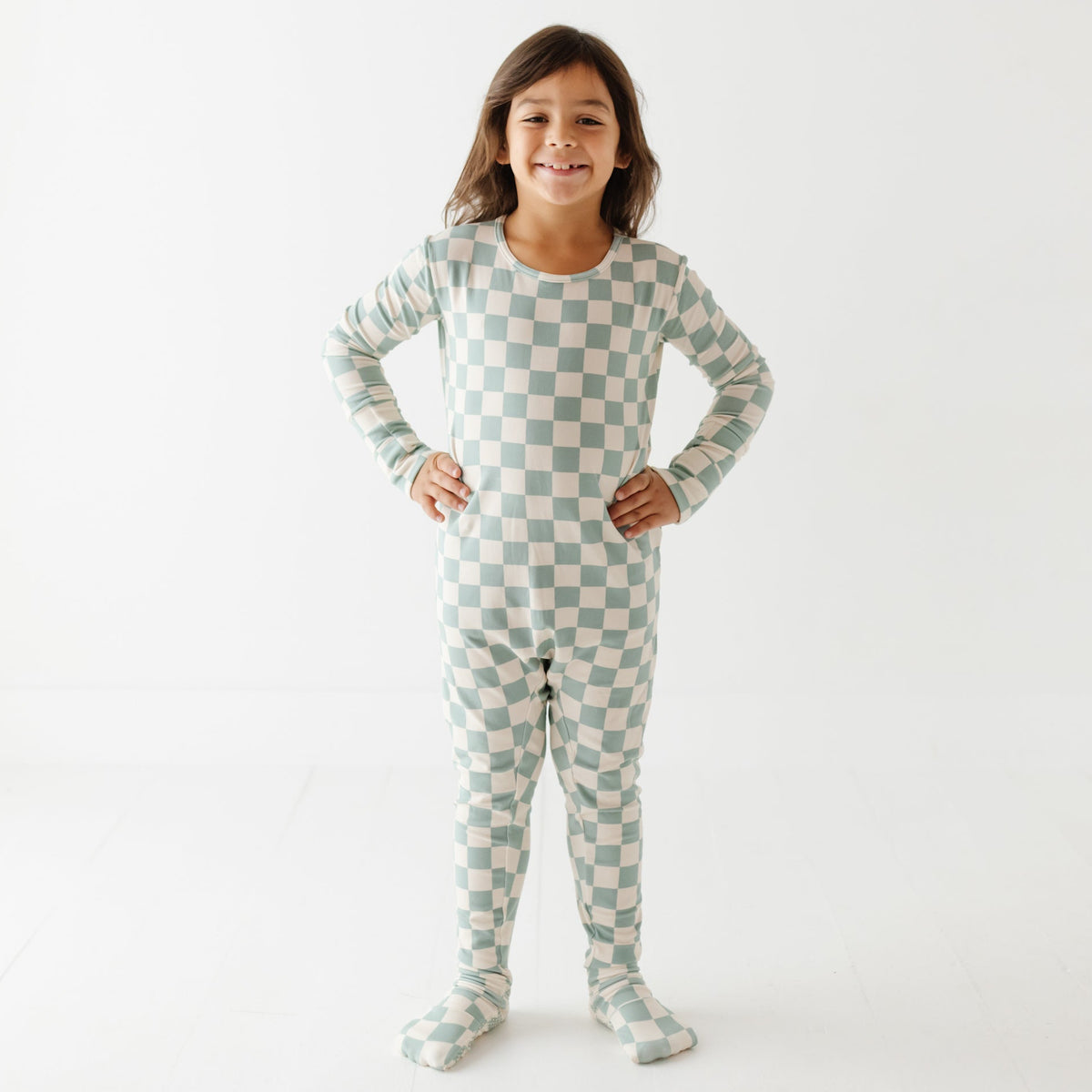 A kid stands on white floor wearing turquoise and white checked romper. 