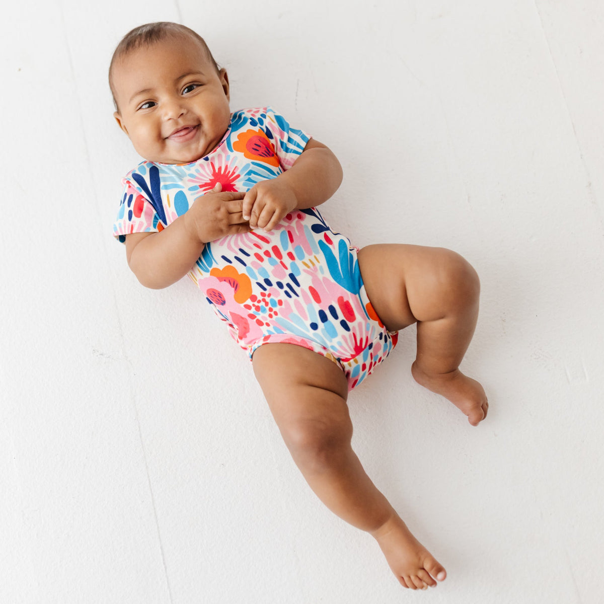 A smiling baby lays on white floor wearing a colorful floral onesie. 