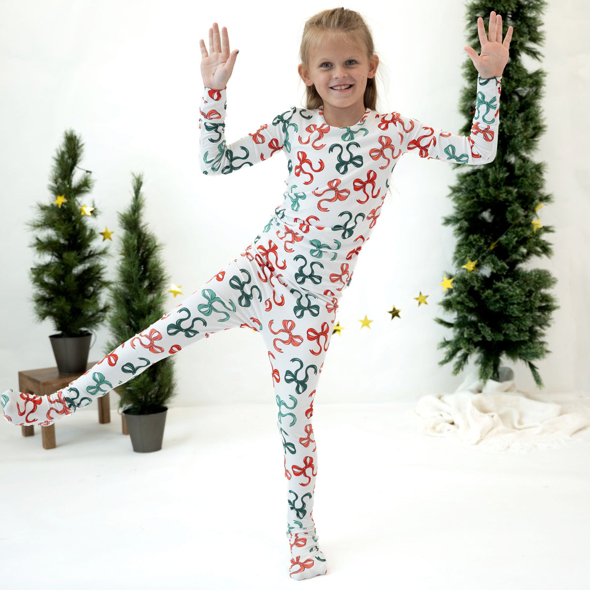 A young girl wearing a long-sleeve, full-length white footed pajama with a colorful repeating bow pattern, posing in a festive studio decorated with small potted Christmas trees and gold star decorations.