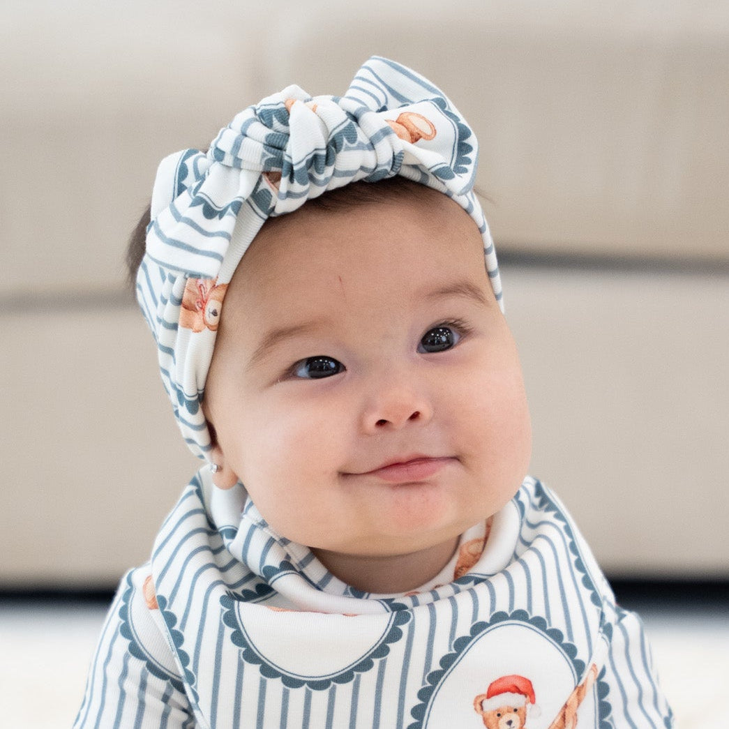 A baby sitting on a white shag rug wearing a matching long-sleeve romper and bow headband set with a blue and white pinstripe pattern and cartoon monkey illustrations.