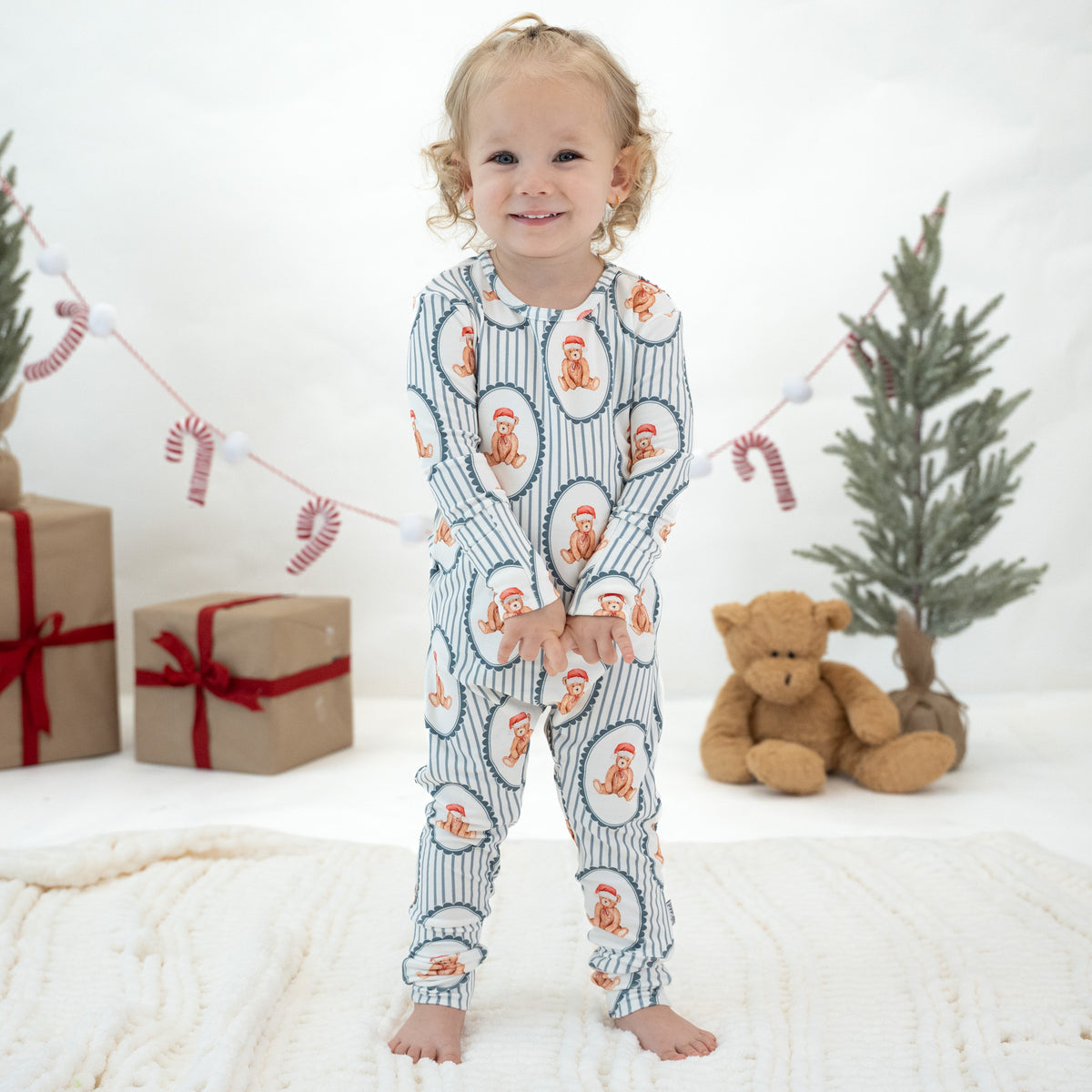 A toddler stands barefoot on a white knit blanket wearing a long-sleeve pajama one-piece with a holiday print. The pajamas have a navy blue and white vertical stripe pattern with circular motifs containing Santa silhouettes and small red accents.