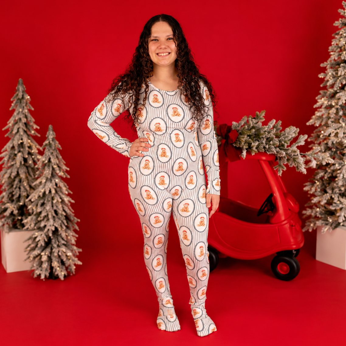 A woman stands in a studio wearing a long-sleeve, full-body footed one-piece pajama with an allover circular fruit print on a white base. She is posed in front of a bright red backdrop with decorative flocked Christmas trees and a red toy car.