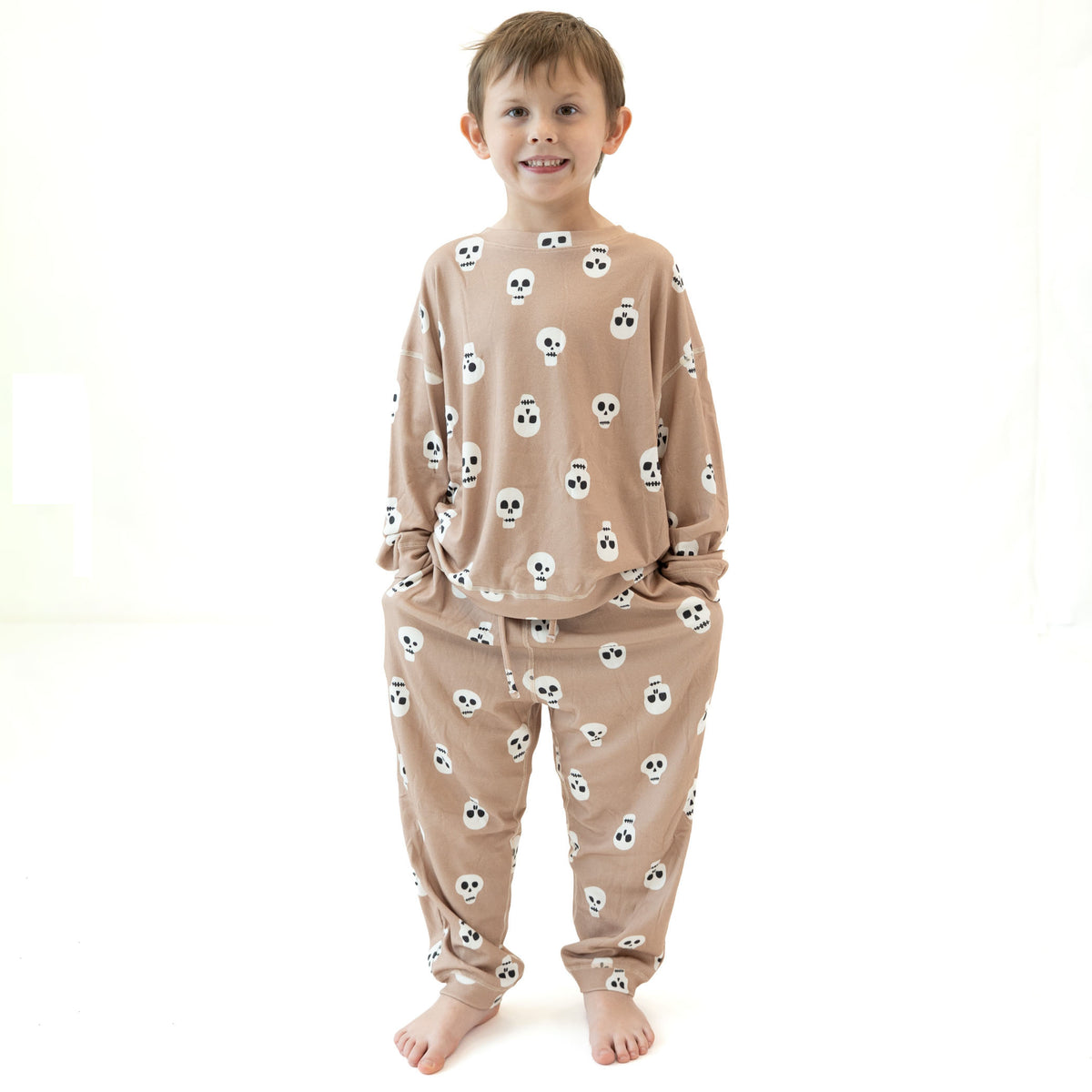 A young boy wearing a beige long-sleeve pajama set with a white skull pattern, standing against a bright white background.