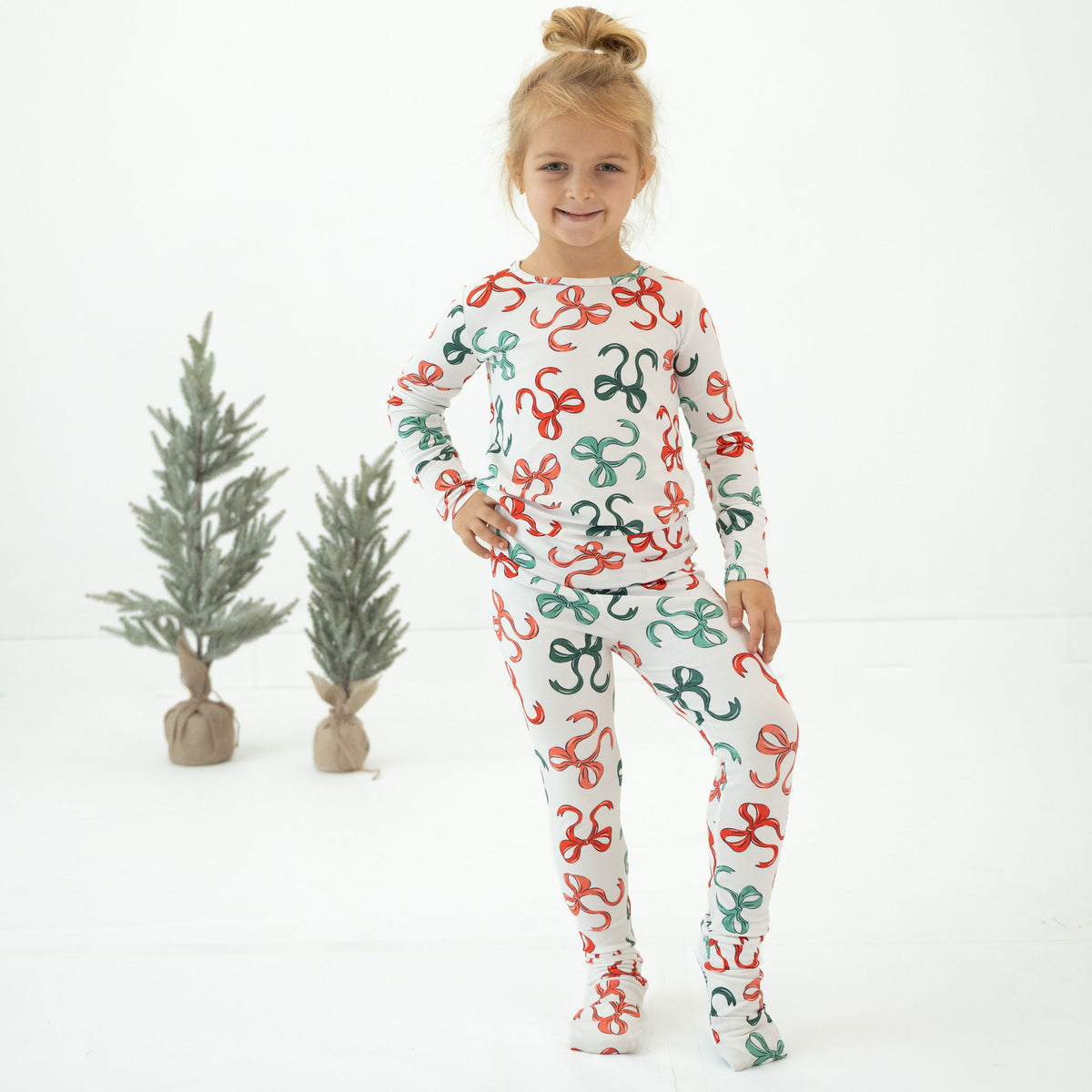 A young girl with blonde hair in a bun stands wearing a matching long-sleeve pajama set with a festive red and green pattern on a white background. She is posed in front of two small potted artificial pine trees against a seamless white studio backdrop.
