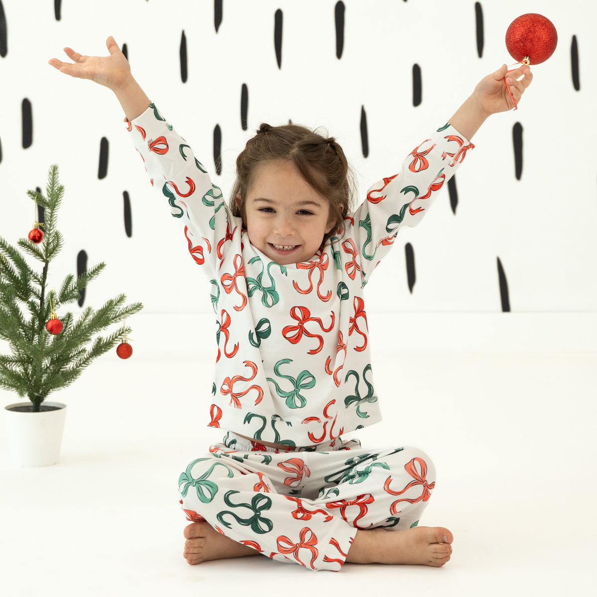 A young girl sits cross-legged on a white floor, wearing a matching white long-sleeve pajama set with a playful red and green abstract print. She is holding a red ornament, with a small decorated Christmas tree and a black-and-white patterned wall in the background.