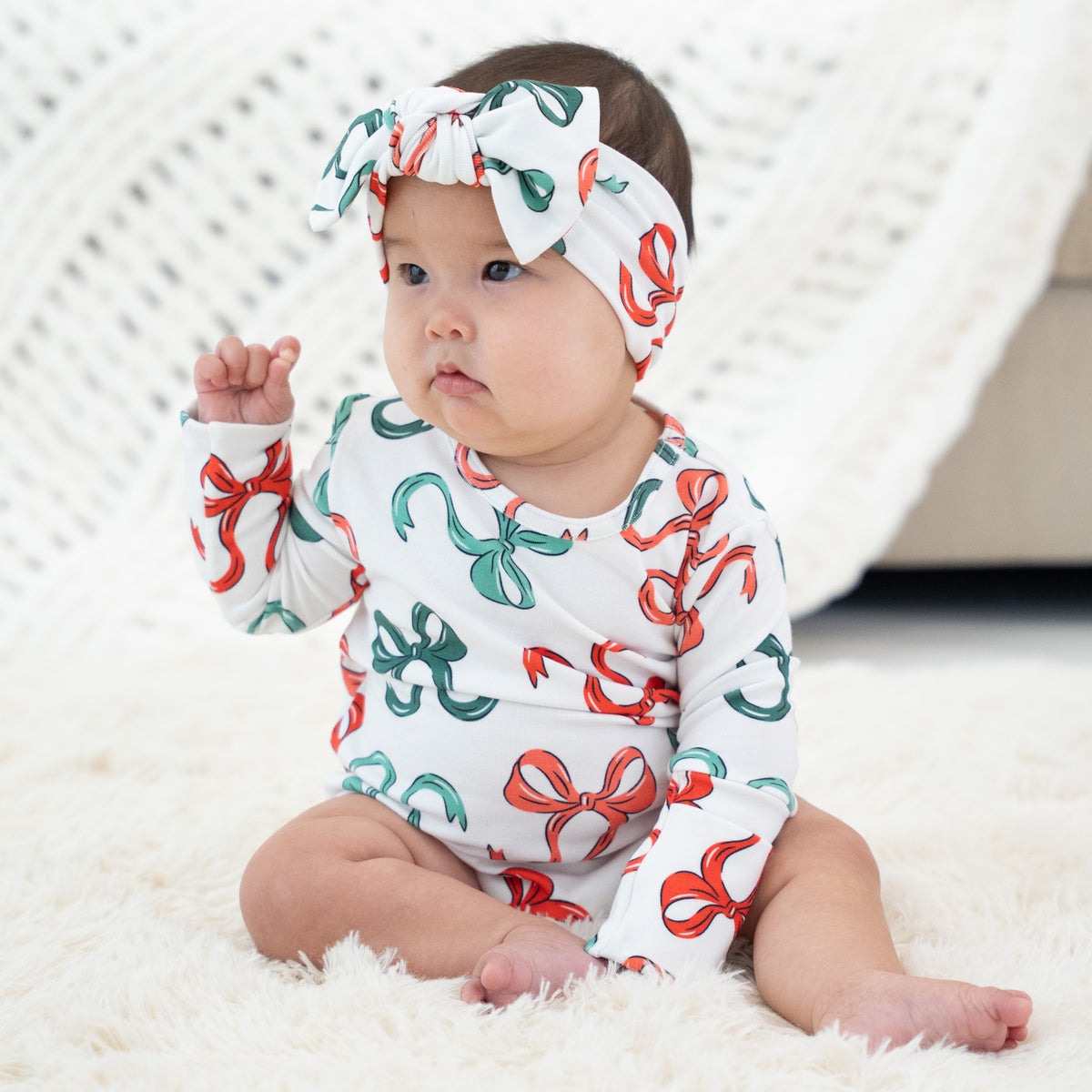 A baby sitting on a white fluffy rug wearing a white long-sleeve romper and matching headband with a red and green bow pattern.
