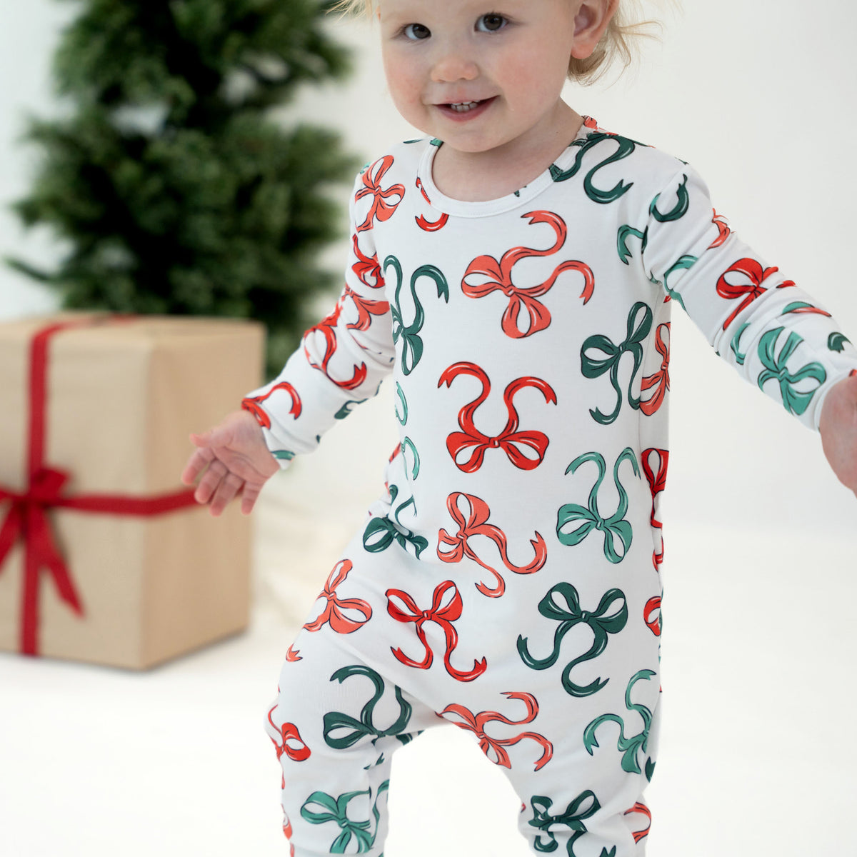 A baby wearing a white long-sleeve footed sleeper with a red and green bow pattern, standing in front of a blurred Christmas tree and a wrapped gift box.