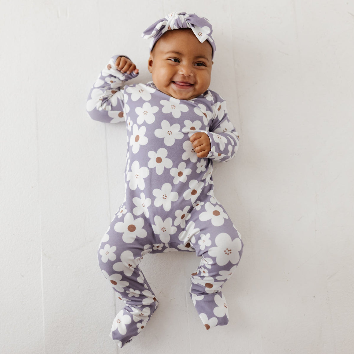 A smiling baby lays on a white floor wearing a lavender romper with white daisies on it. 