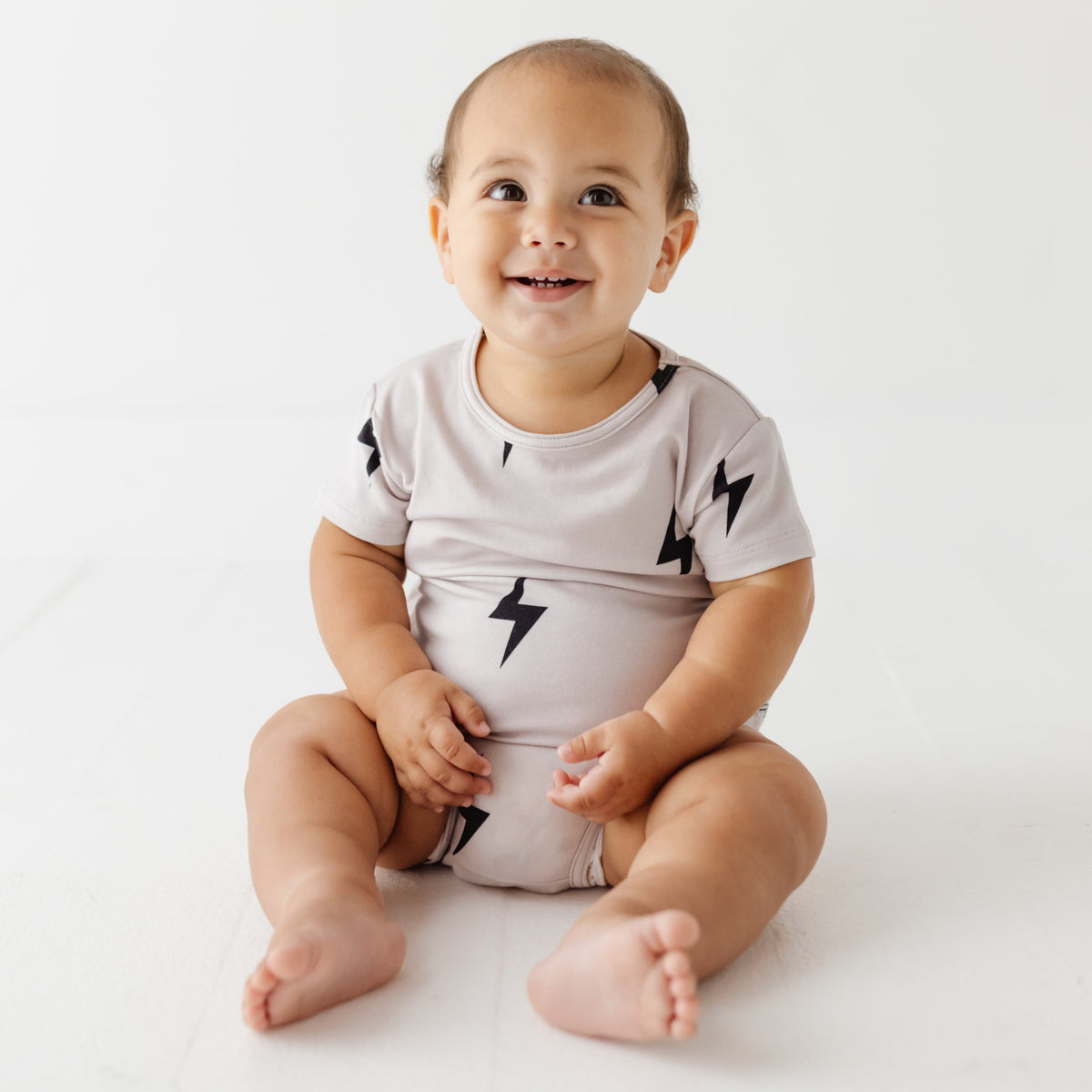 Baby sits on white floor wearing taupe onesie with lightening bolts on it. 