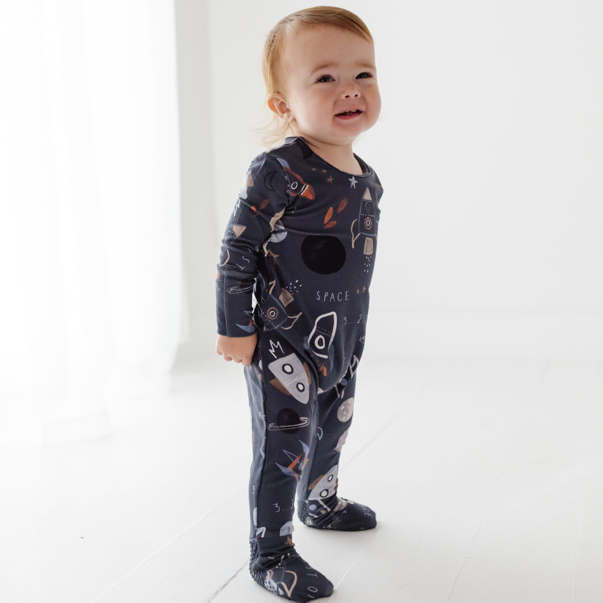 A smiling baby stands on a white floor. Wearing a navy blue romper with a space-themed design on it. 