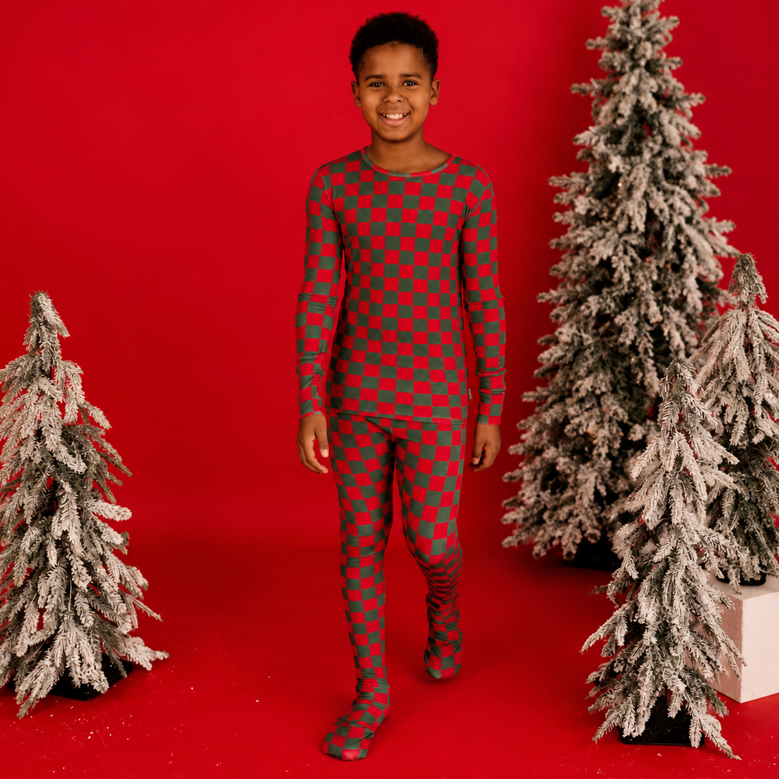 A young boy stands against a bright red studio backdrop wearing a long-sleeve, full-length red and green checkered pajama set. Two artificial frosted Christmas trees are placed on either side of him, with one tree on a white box.