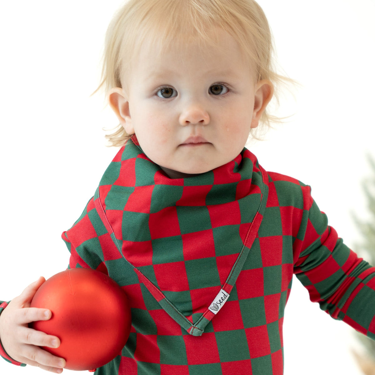 A baby wearing a red and green checkered long-sleeve top and matching bib, holding a red ornament against a white background.