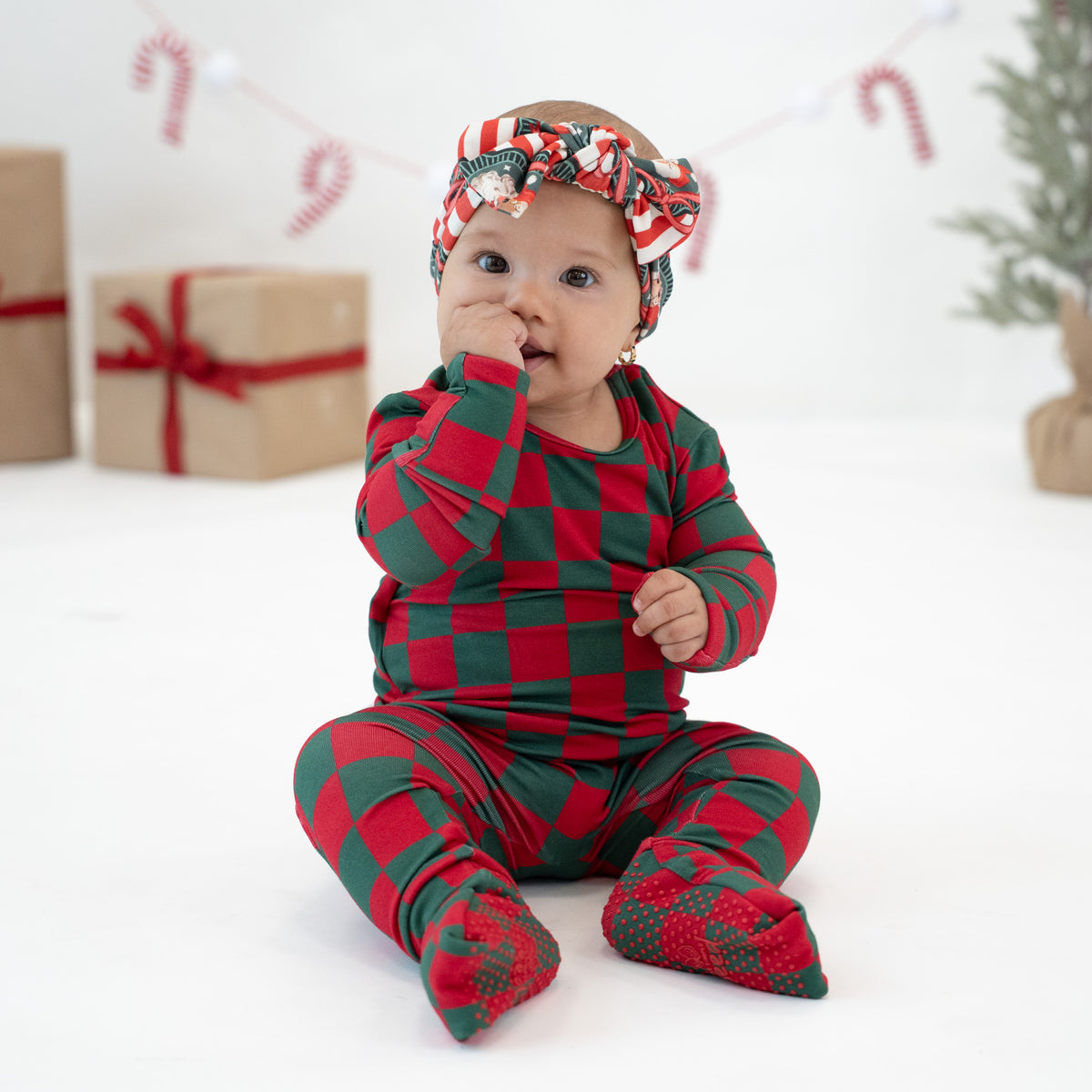 A baby sitting on a white studio floor wearing a red and green checkered long-sleeve footed onesie and a matching red and green checkered headband with a small ornament detail.