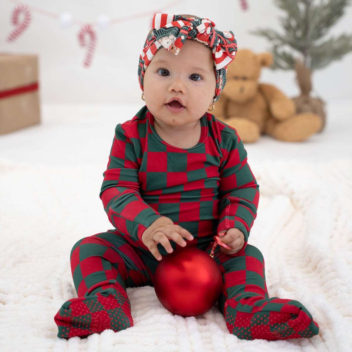 A baby sitting on a white textured blanket wearing a red and green checkered holiday footie pajama set and a matching headband, holding a red ornament with a teddy bear and candy cane decorations in the background.