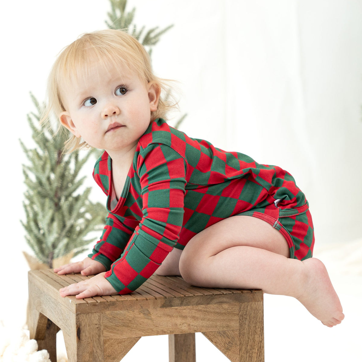 A baby wearing a red and green checkered long-sleeve romper sits on a small wooden stool in a bright, clean indoor setting.