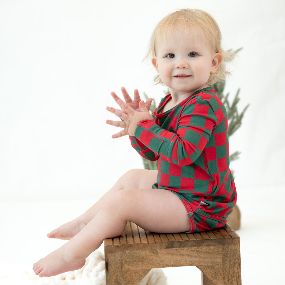 A toddler girl with blonde hair sits on a wooden stool wearing a red and green checkered long-sleeve romper. She is clapping her hands against a clean white studio background.
