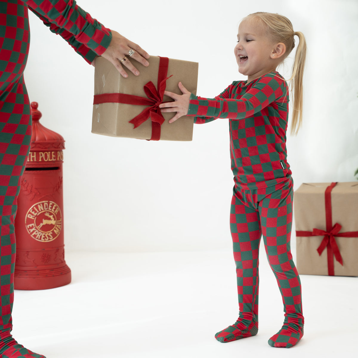 A young girl in a red and green checkered pajama set reaches for a wrapped gift box handed to her by an adult, with a red vintage-style mailbox prop on a white studio background.