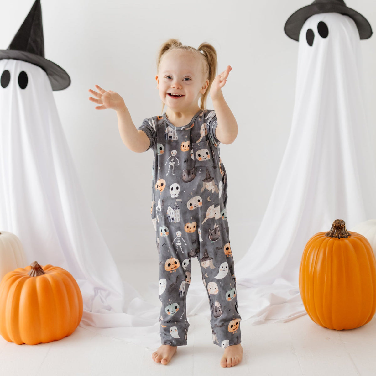 A smiling girl stand on white floor with ghost and pumpkins in the background. She is wearing a great romper with a Halloween-themed design. 