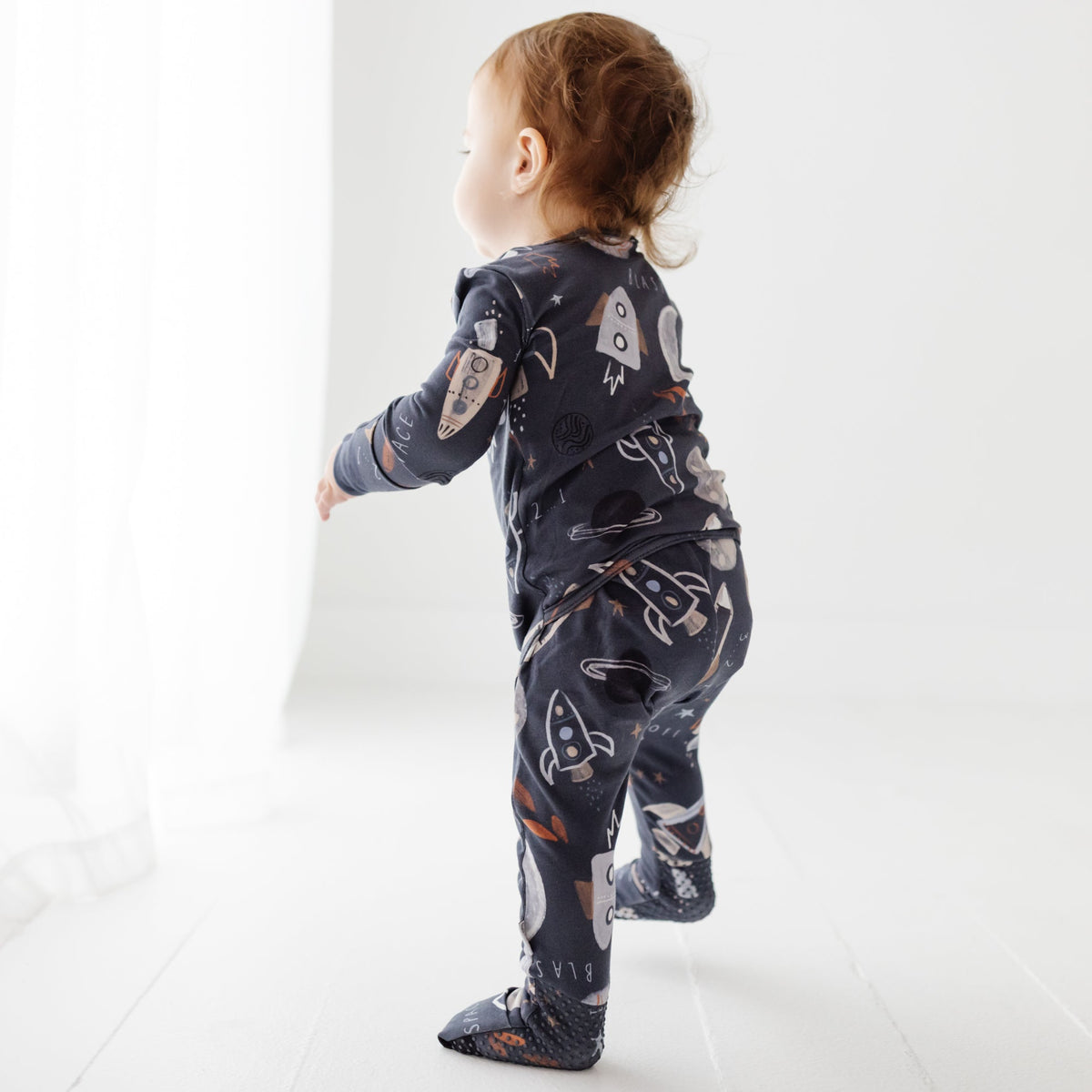 A baby stands on a white floor. Wearing a navy blue romper with a space-themed design on it. 