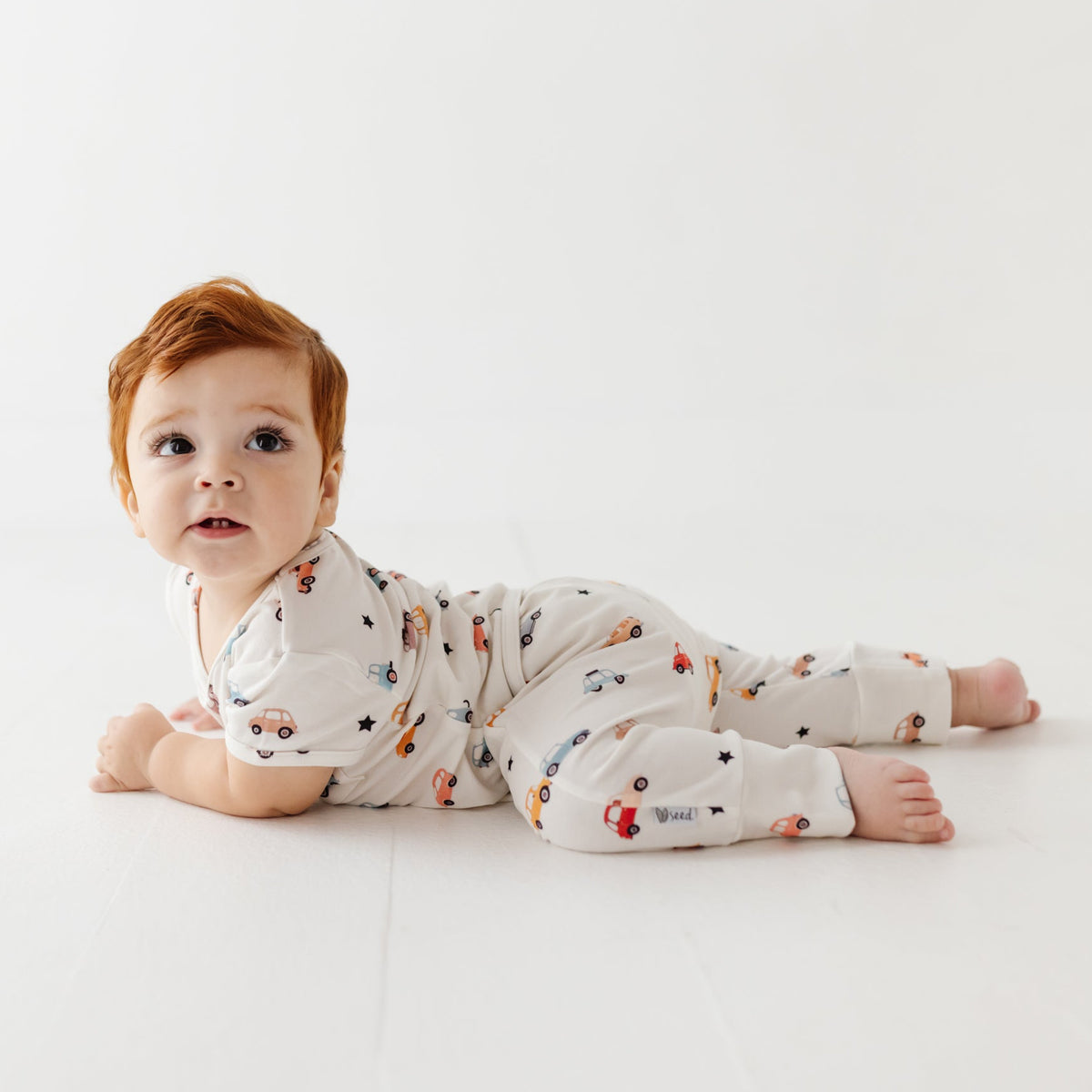 A baby lays on white floor wearing white romper with car print. 
