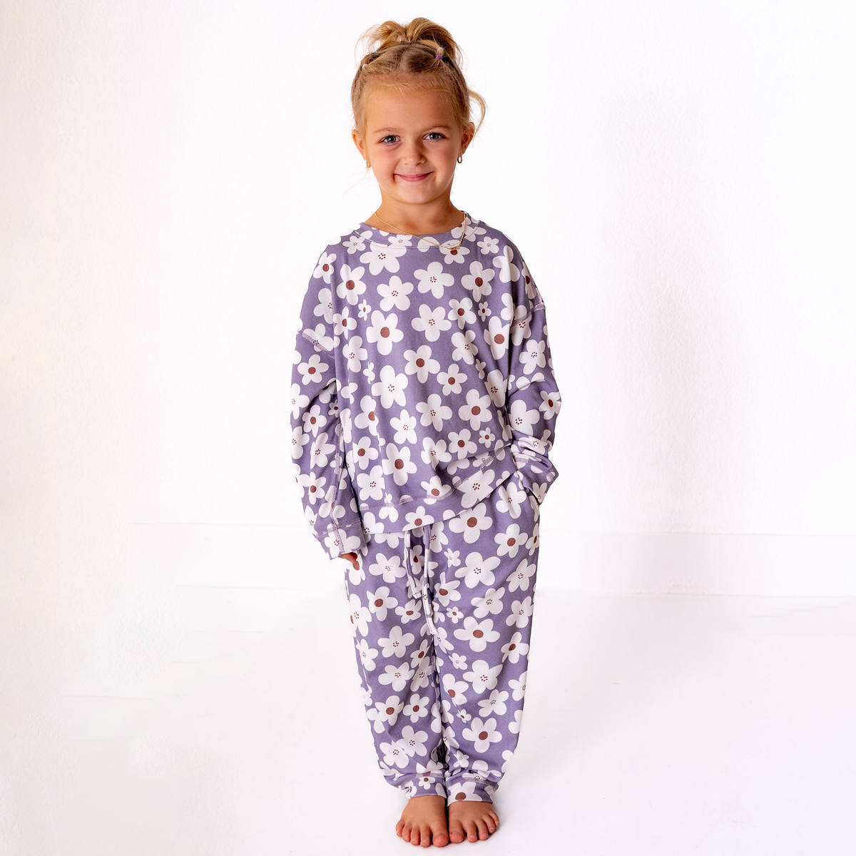 A young girl with blonde hair in a bun stands barefoot against a white studio background, wearing a matching long-sleeve pajama set with a purple base and a white daisy pattern with small orange centers.