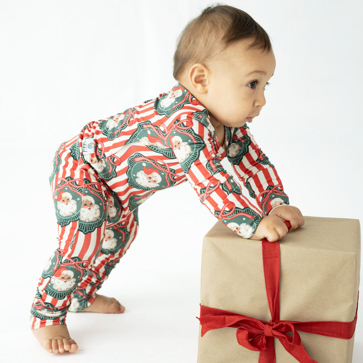 A baby wearing red and white striped Christmas-themed pajamas with Santa Claus patterns, crawling while pushing a brown gift box tied with a red ribbon against a white background.