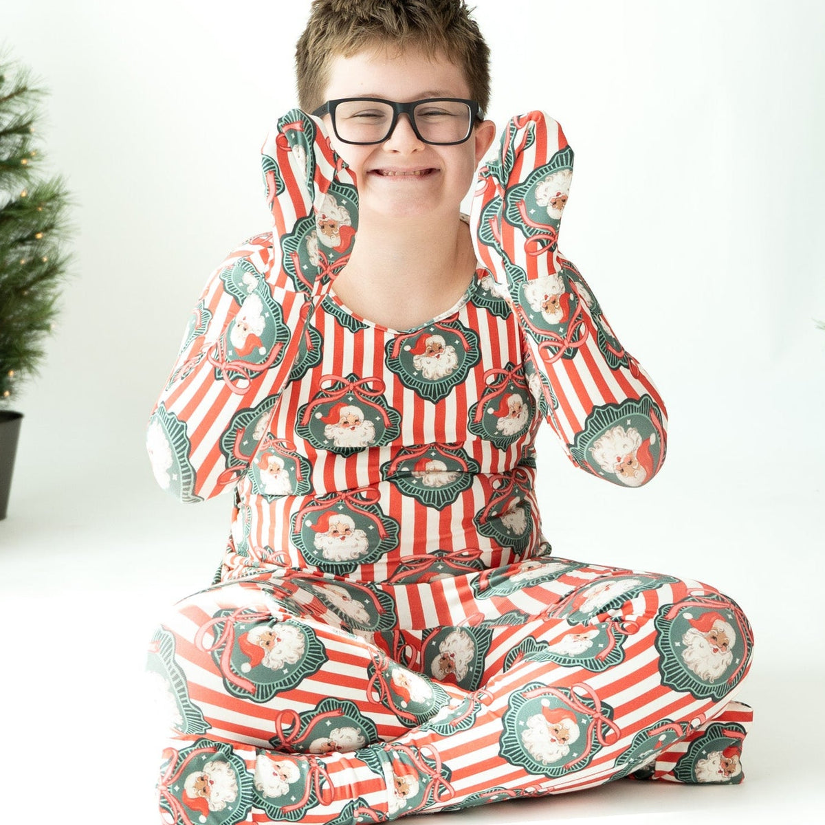 A child wearing a red and white striped Christmas-themed pajama set with green wreath patterns sits cross-legged on a white studio floor, surrounded by holiday decorations including a red post box and Christmas trees.