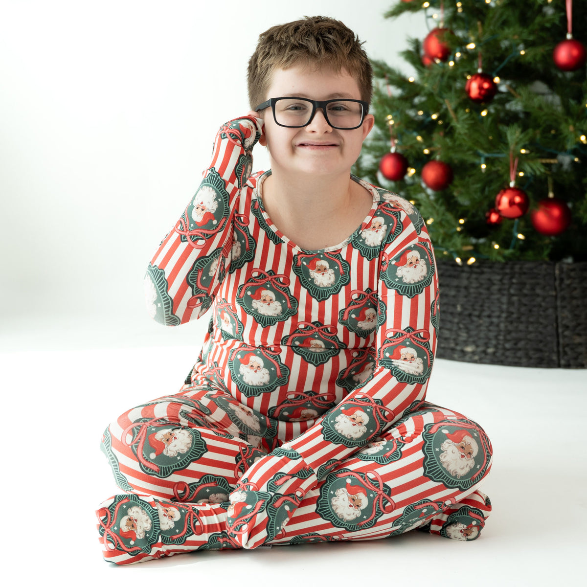 A child wearing red and white striped Christmas pajamas with Santa Claus face prints, sitting cross-legged on a white floor next to a decorated Christmas tree.