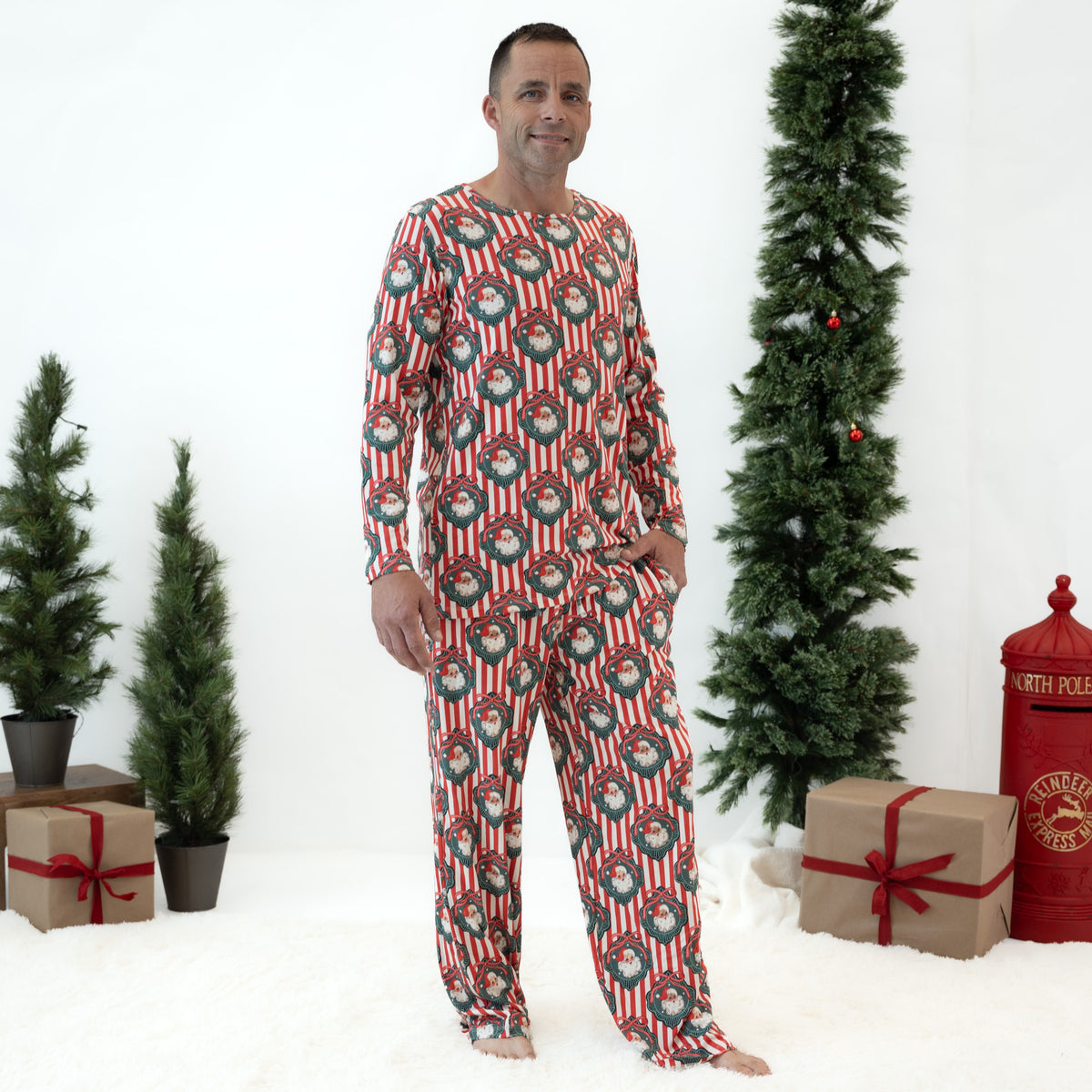 A man wearing a festive Christmas-themed long-sleeve pajama set with a red, white, and green pattern, standing in a studio decorated with artificial Christmas trees and wrapped gifts.