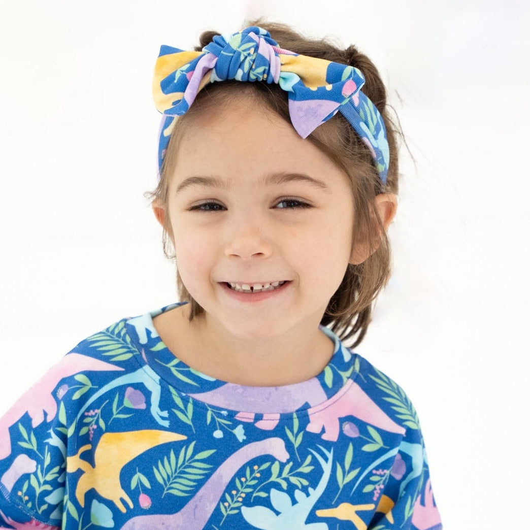 A young child sitting cross-legged on a white studio background, wearing a matching long-sleeve pajama set and headband with a colorful dinosaur print on a blue base.