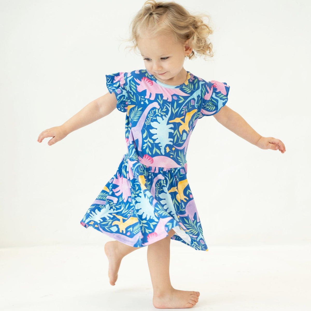 A toddler girl with blonde hair in a ponytail is wearing a short-sleeve dress with a colorful dinosaur and tropical leaf print, captured mid-step against a plain white studio background.