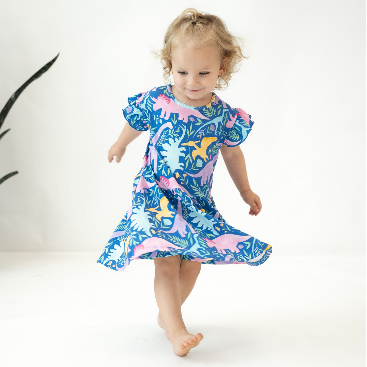 A toddler girl with blonde curly hair is wearing a short-sleeve dress with a colorful dinosaur print, walking barefoot on a white studio floor.