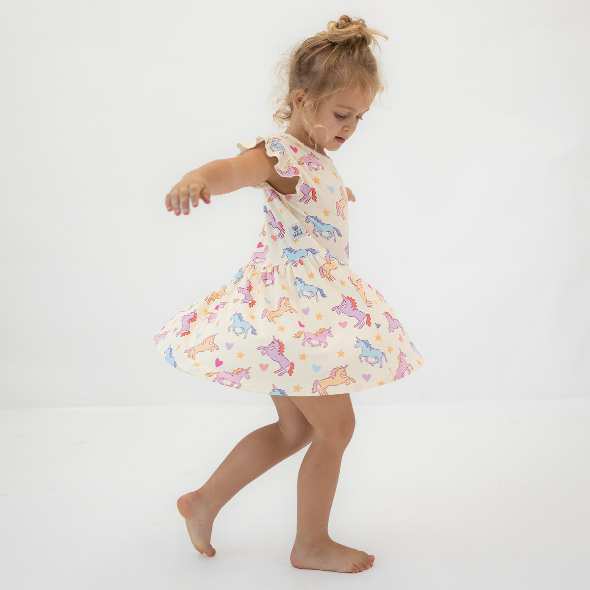 A young girl with blonde hair in a bun is wearing a sleeveless cream-colored dress with a colorful unicorn print, captured mid-spin against a plain white studio background.