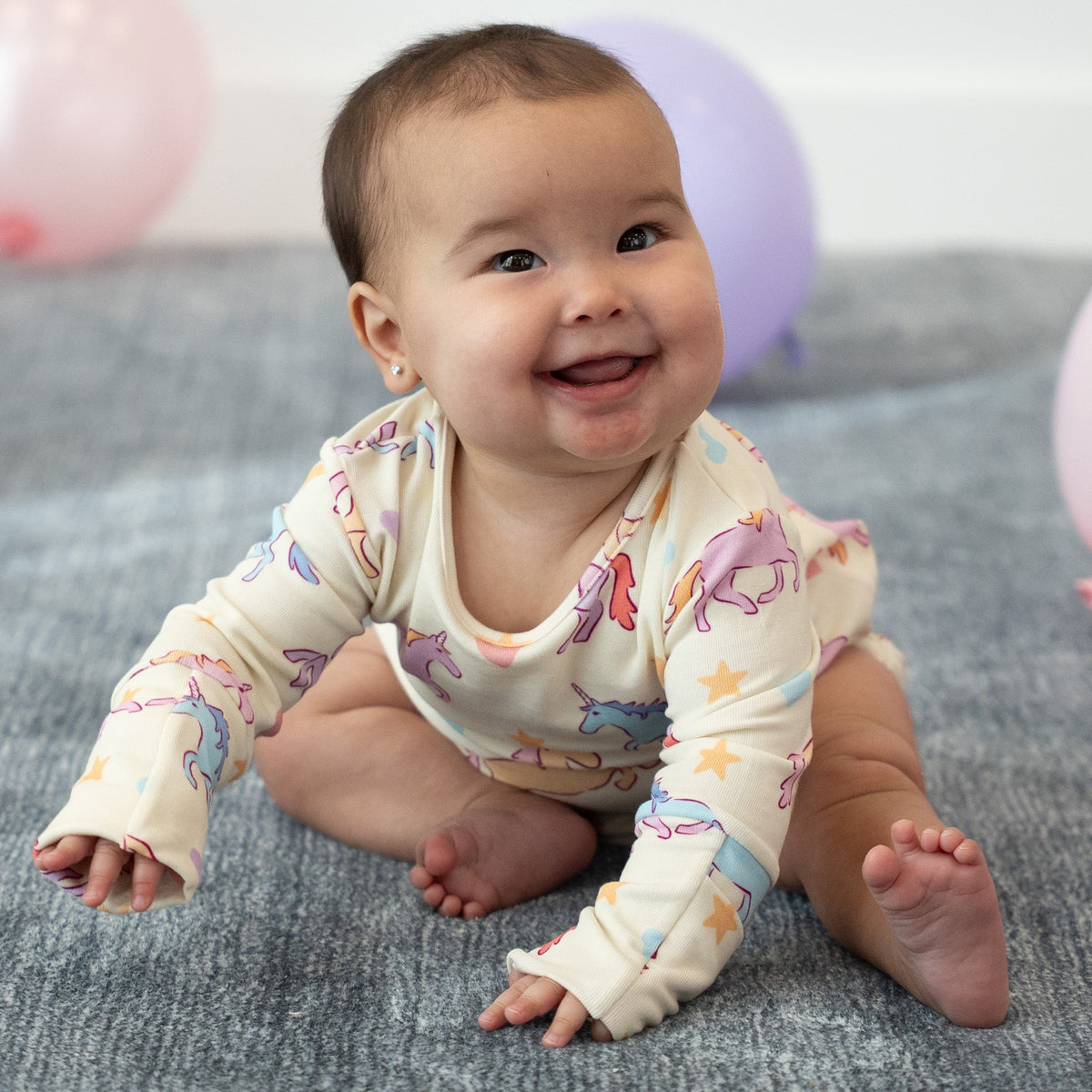 A smiling baby sitting on a gray blanket wearing a cream long-sleeve bodysuit with a colorful unicorn and star print, with pink and purple balloons in the background.