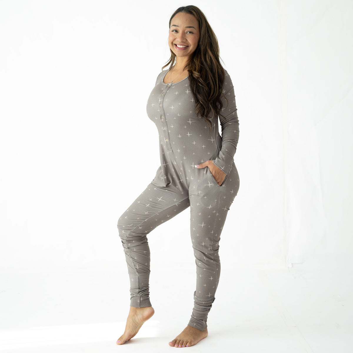 A woman modeling a light gray long-sleeve zip-up pajama one-piece with a small white star pattern, standing against a white studio background.