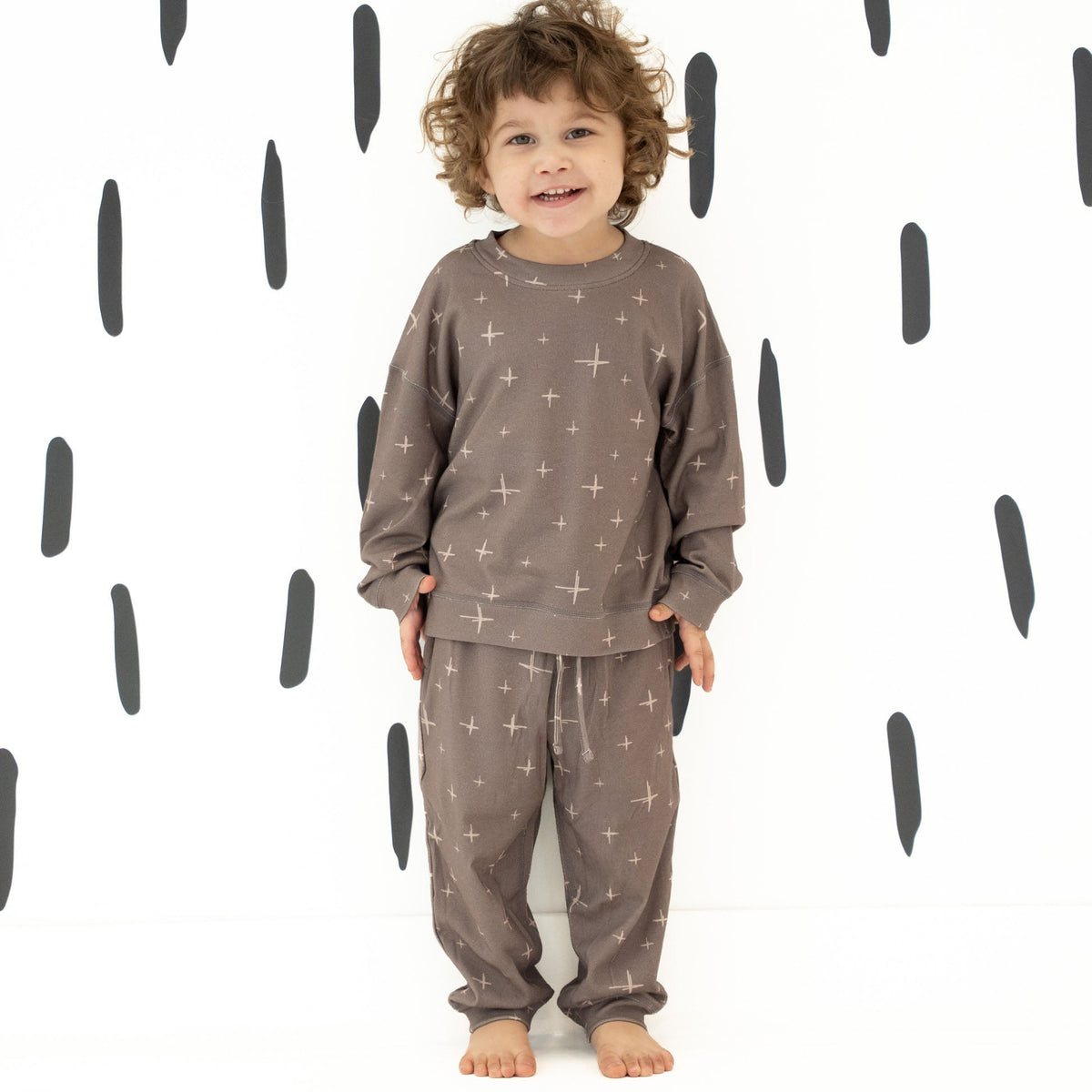A toddler stands barefoot against a white wall with black abstract vertical marks, wearing a matching two-piece pajama set in taupe-brown with a light star pattern.