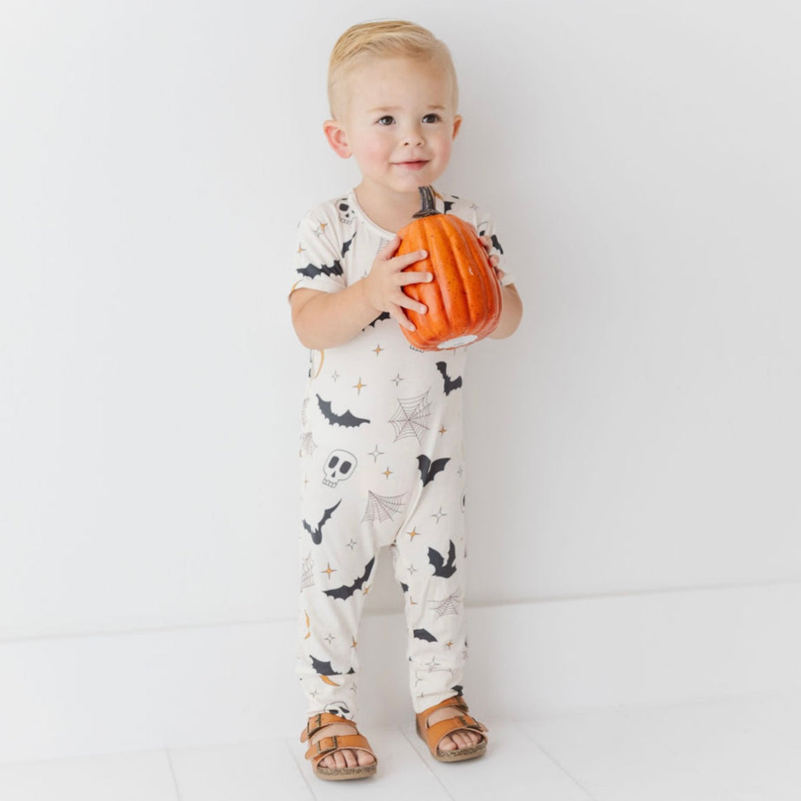Child wearing a Halloween-themed onesie holding a small pumpkin against a white background
