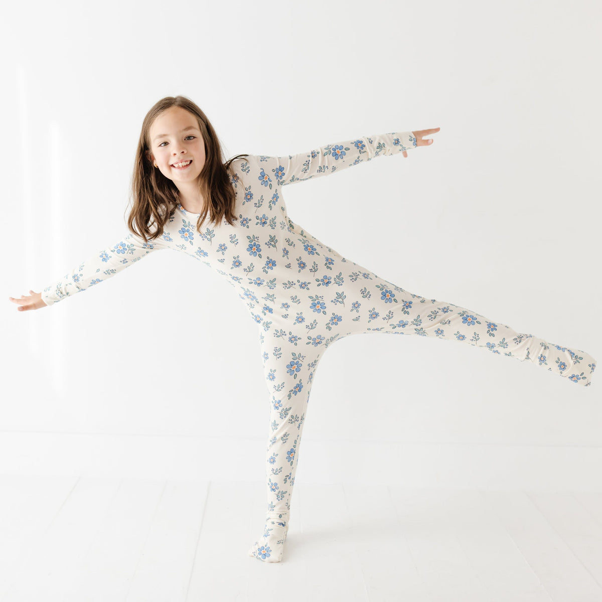 A girl stands with her arms out on a white floor wearing a cream rompers with blue flowers. 