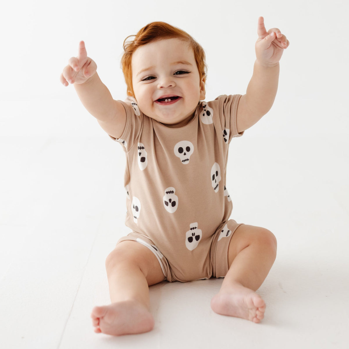 A smiling baby sits on white floor wearing tan romper with skulls on it. 