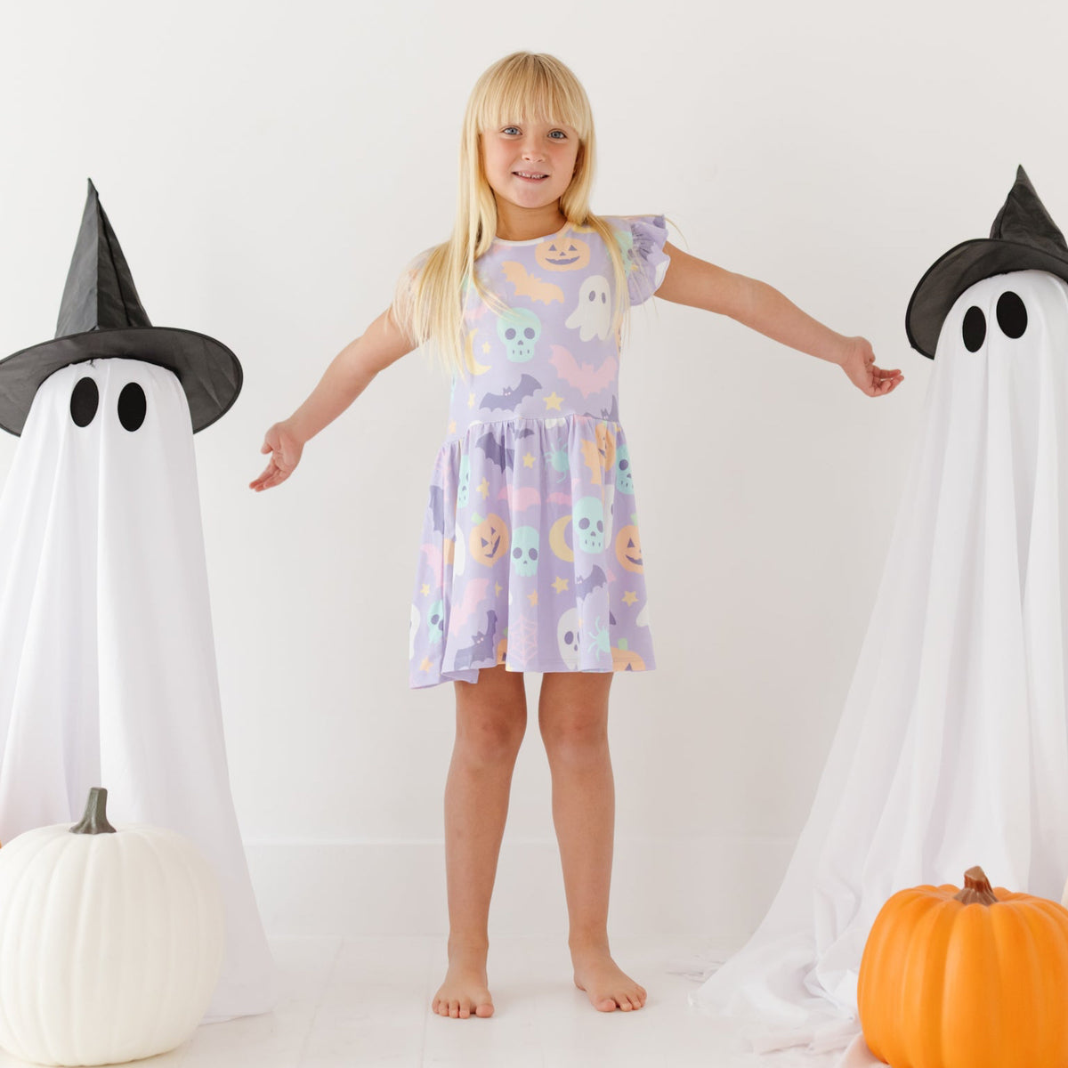 A smiling girl stand on a white floor with pumpkins and ghosts in the background. She is wearing a lavender dress and shorts with a Halloween-themed print.