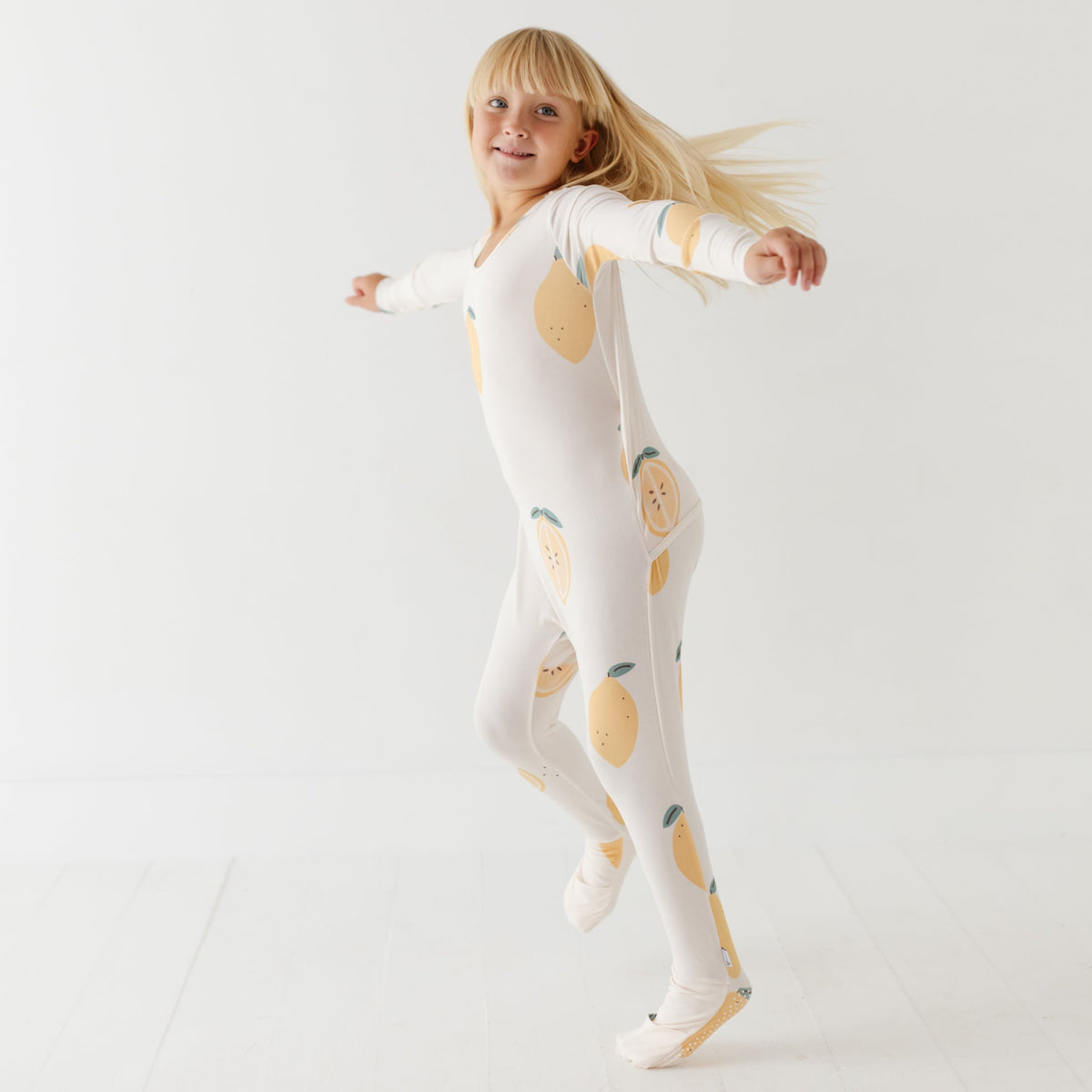 A smiling girl stand on white floor wearing a cream rompers with lemons on it. 