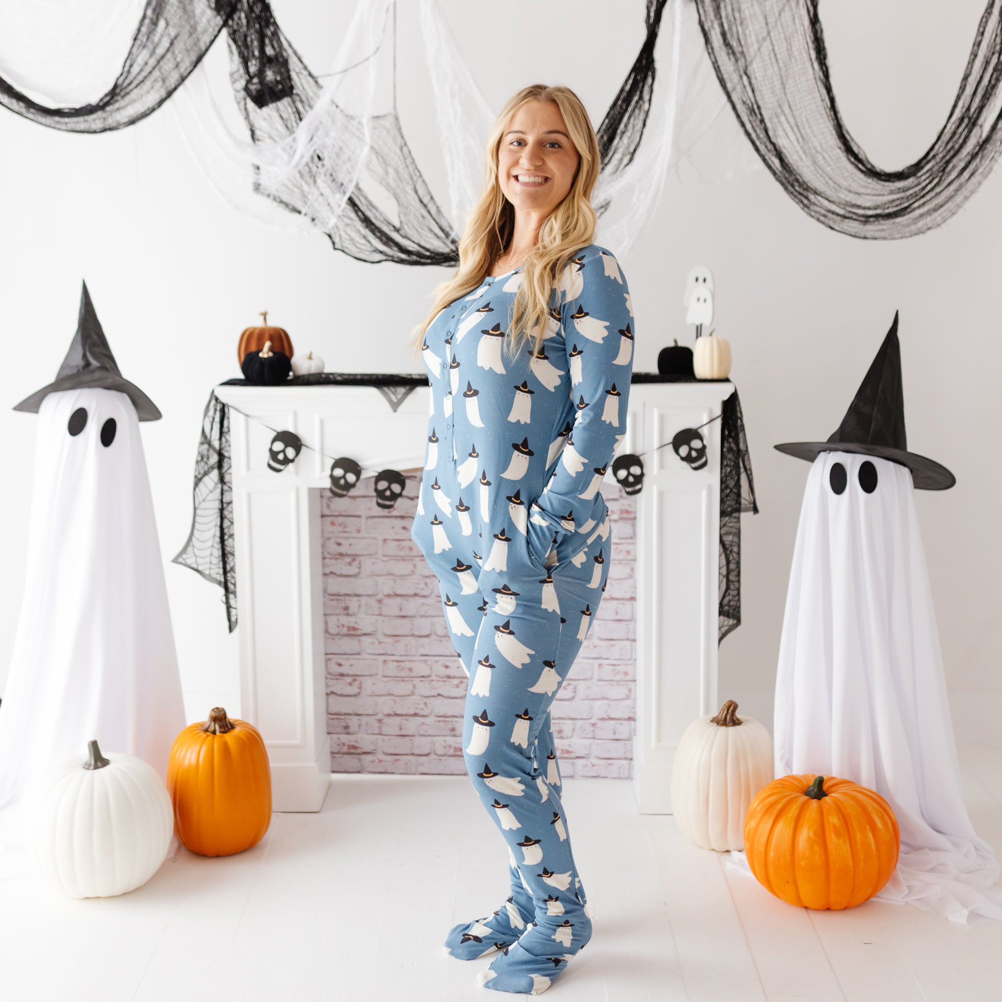 A woman stands in a Halloween-themed room wearing a light blue long-sleeve pajama set with a white ghost pattern. The room features a white fireplace mantel decorated with black spiderwebs, black witch hats, skull garlands, and pumpkins, with two large ghost decorations on either side of the fireplace.