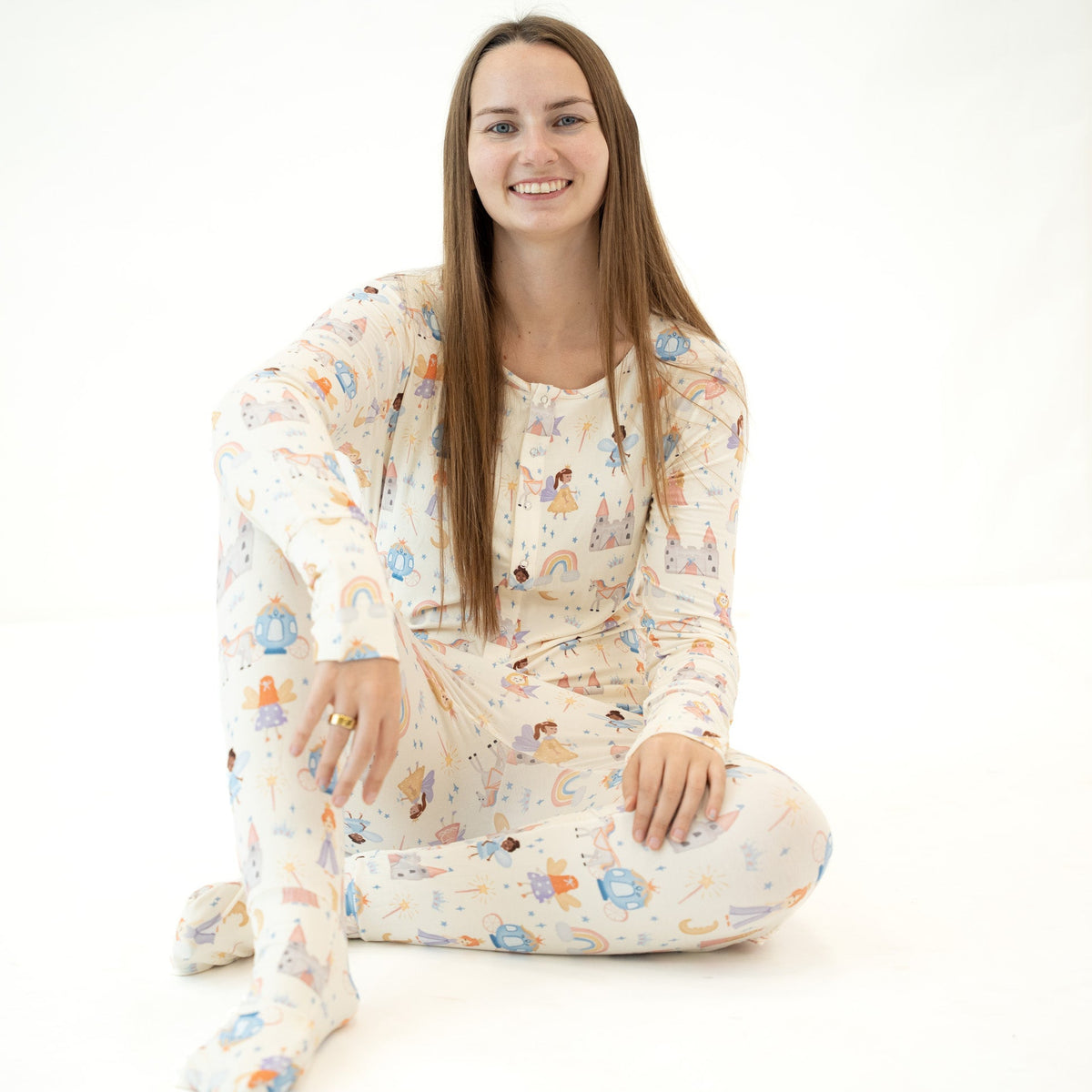 A smiling young woman with long light brown hair sits cross-legged on a seamless white studio background, wearing a matching long-sleeve pajama set with a whimsical multicolor cartoon print on an off-white base.